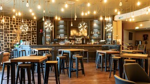 Un restaurante con varias mesas, taburetes de bar, una zona de bar con estantes llenos de botellas y lámparas colgantes.