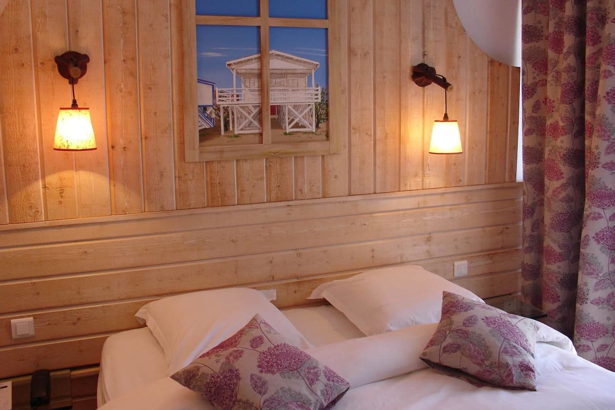 Hotelkamer met een houten hoofdbord, een bed met witte beddengoed en bloemenkussens, twee wandlampen en een raam met uitzicht op een gebouw.