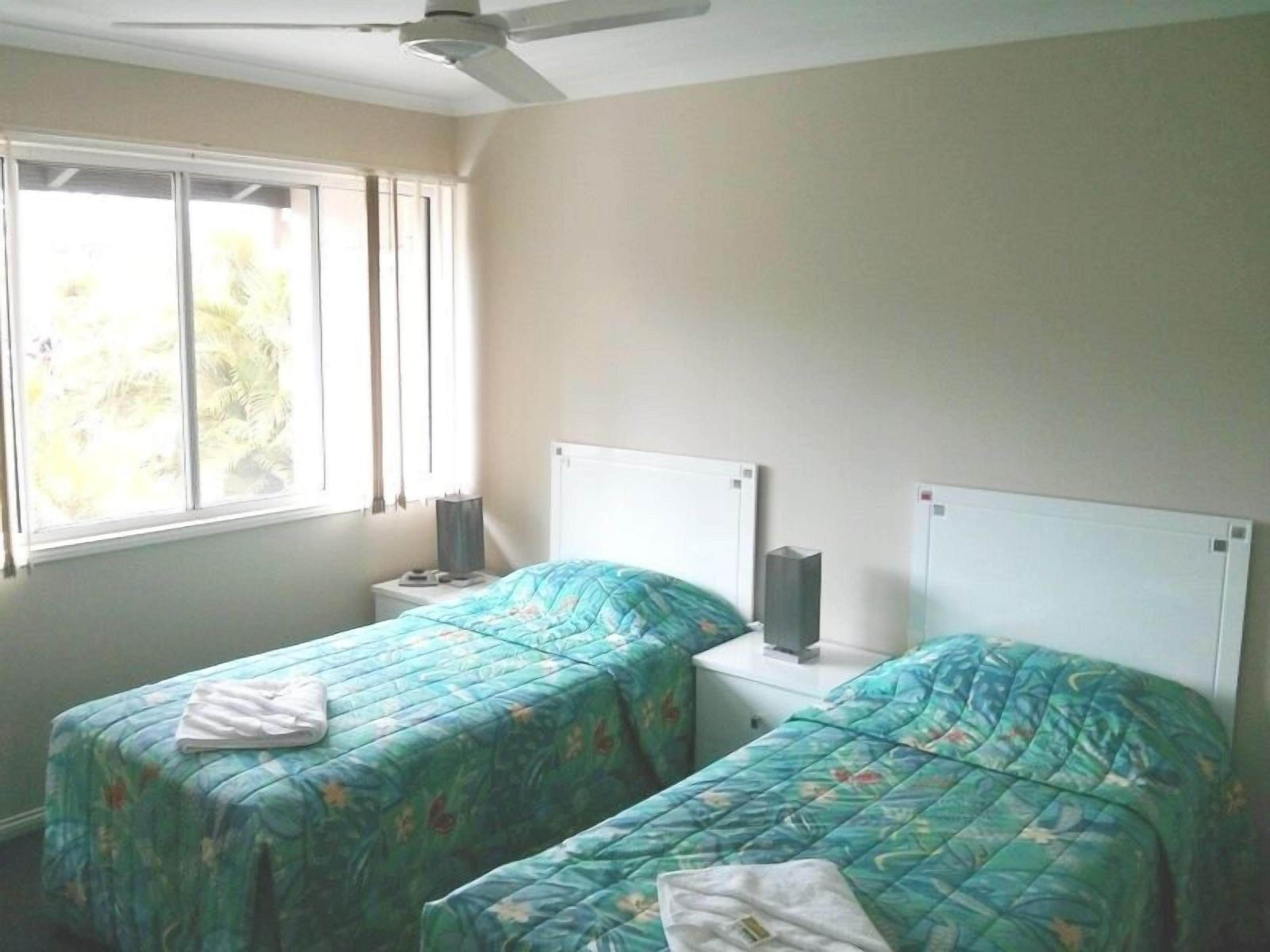 A bedroom with two single beds, a ceiling fan, a window with blinds, and a bedside table with a lamp.