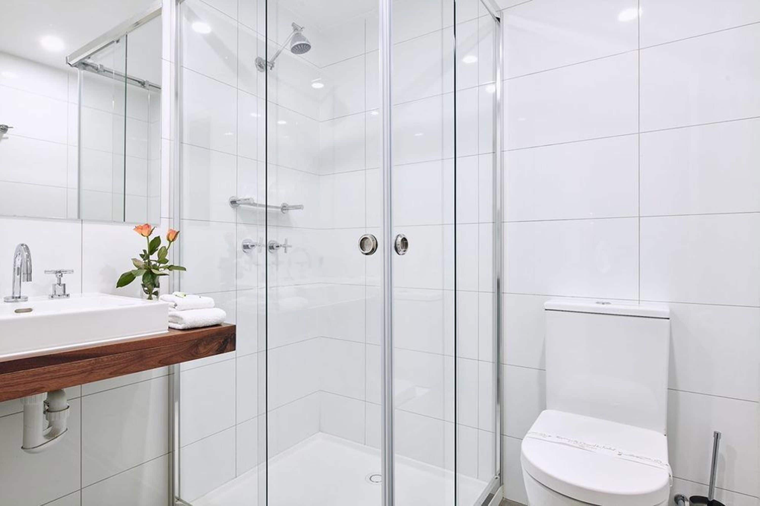 Un baño moderno con ducha envuelta en vidrio, inodoro blanco y tocador de madera con lavamanos y un jarrón de flores.