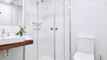 Un baño moderno con ducha envuelta en vidrio, inodoro blanco y tocador de madera con lavamanos y un jarrón de flores.