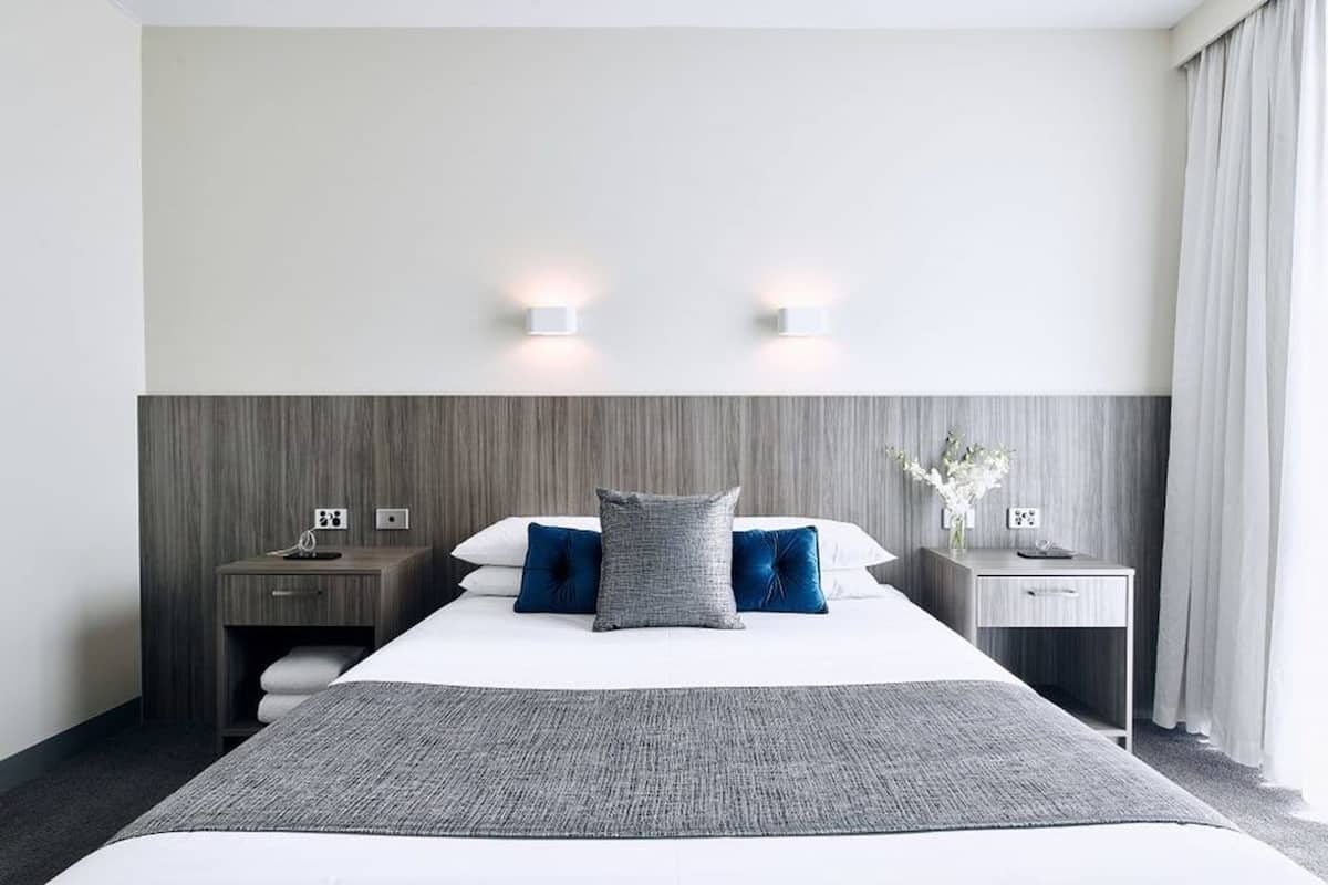 A hotel room with a large bed, two bedside tables, a nightstand, a small desk, and a wall-mounted light fixture.