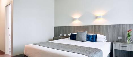 Une chambre d’hôtel avec un grand lit, deux tables de chevet, une petite table et un luminaire fixé au mur.