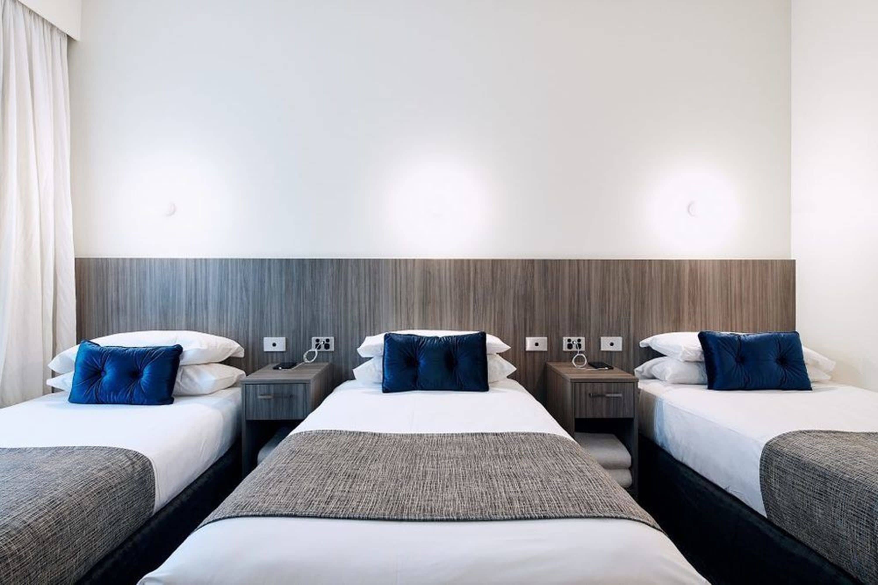 Habitación de hotel con tres camas individuales, cada una con una almohada y una mesita de noche.