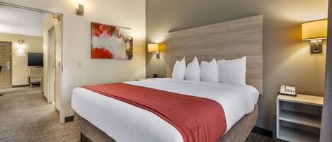 A hotel room with a large bed, a red throw blanket, a headboard with a geometric pattern, a painting on the wall, and a desk with a lamp and phone.
