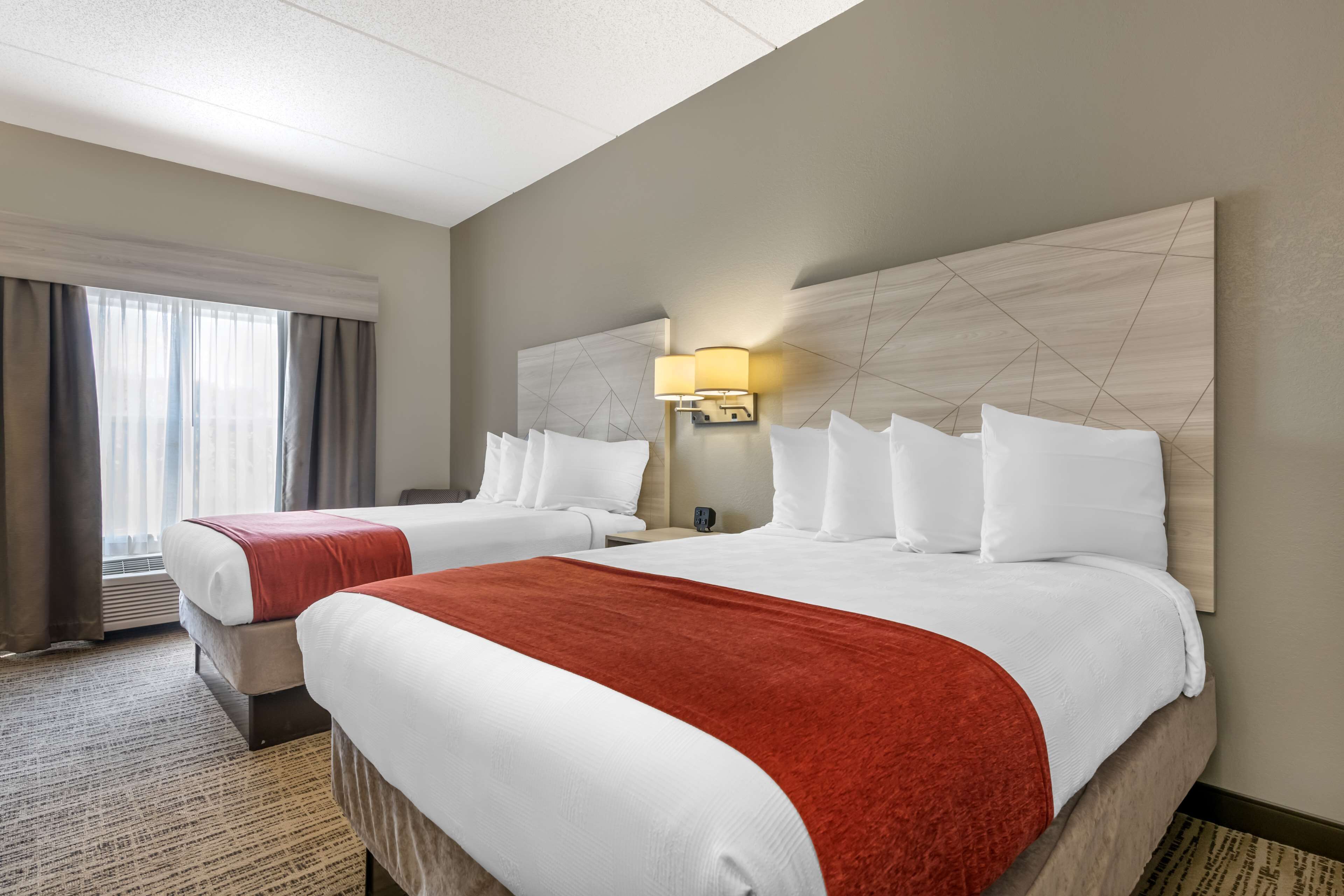 A hotel room with two beds, a large window with curtains, a wall-mounted lamp, and a patterned carpet.