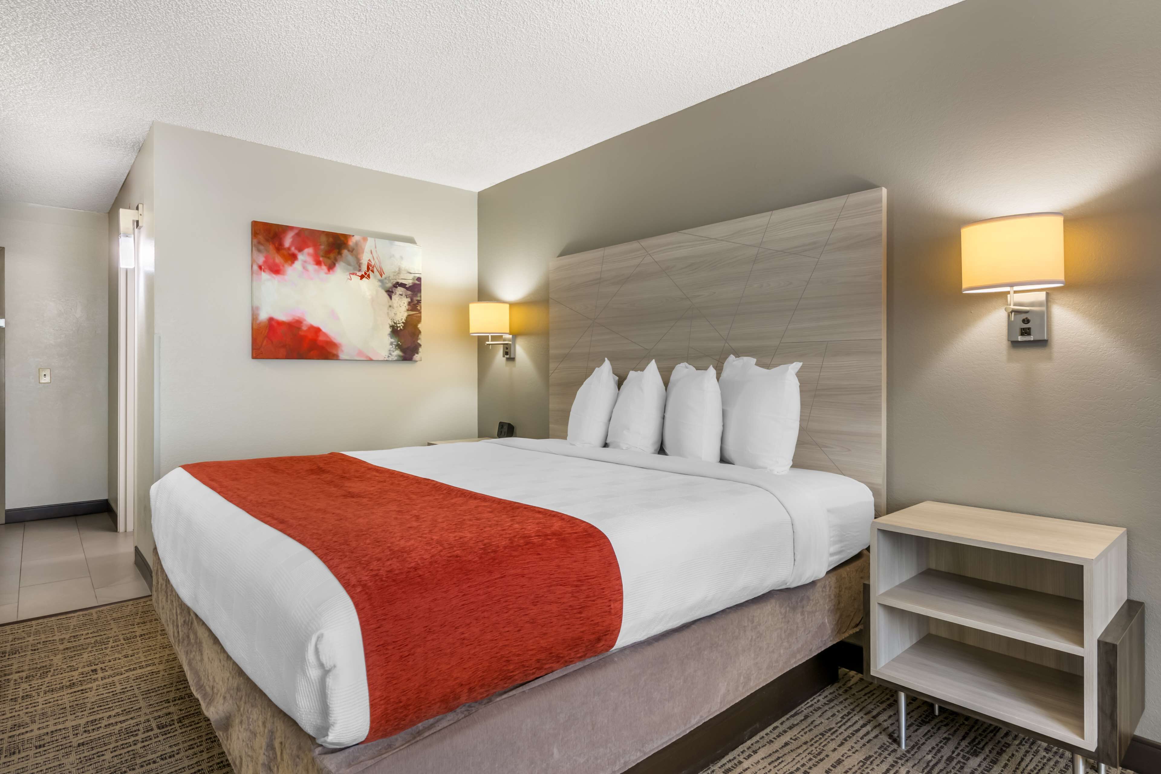Quarto de hotel com uma cama grande, manta vermelha, luminária na parede e um quadro.