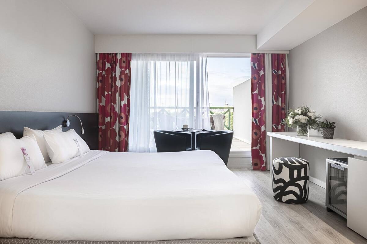 Une chambre d’hôtel avec un grand lit, une petite table, une chaise, un bureau et un balcon avec des rideaux.