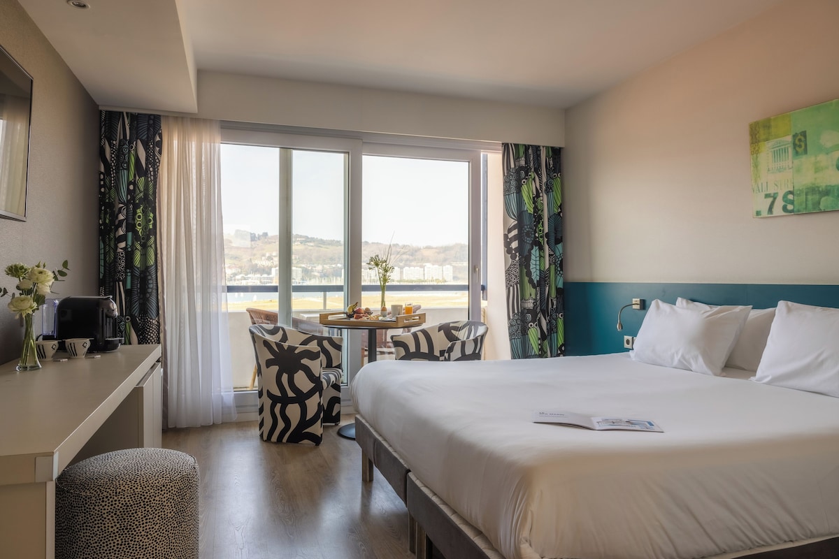 Ein Hotelzimmer mit einem großen Bett, einem Schreibtisch, einem Sessel und Blick auf die Stadt durch das Fenster.