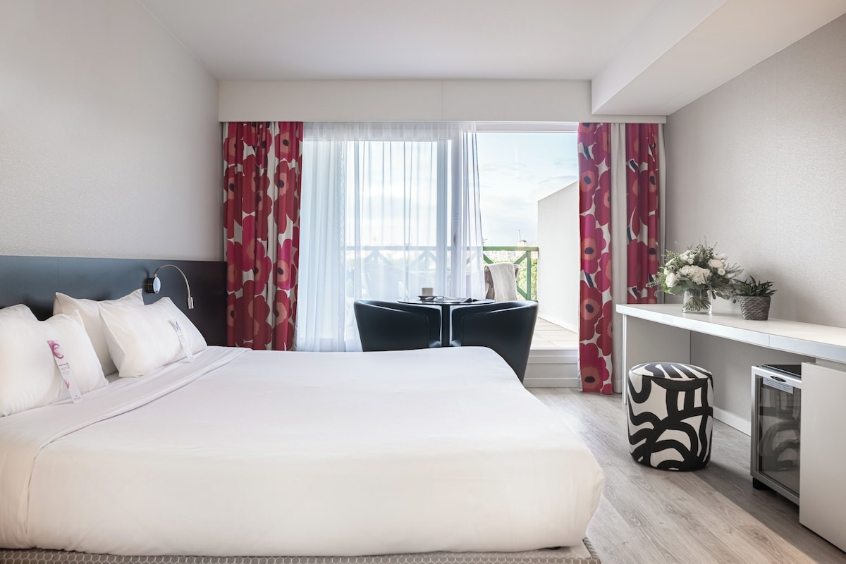 Een hotelkamer met een bed, een bureau, een stoel, een klein tafeltje en een balkon met gordijnen.
