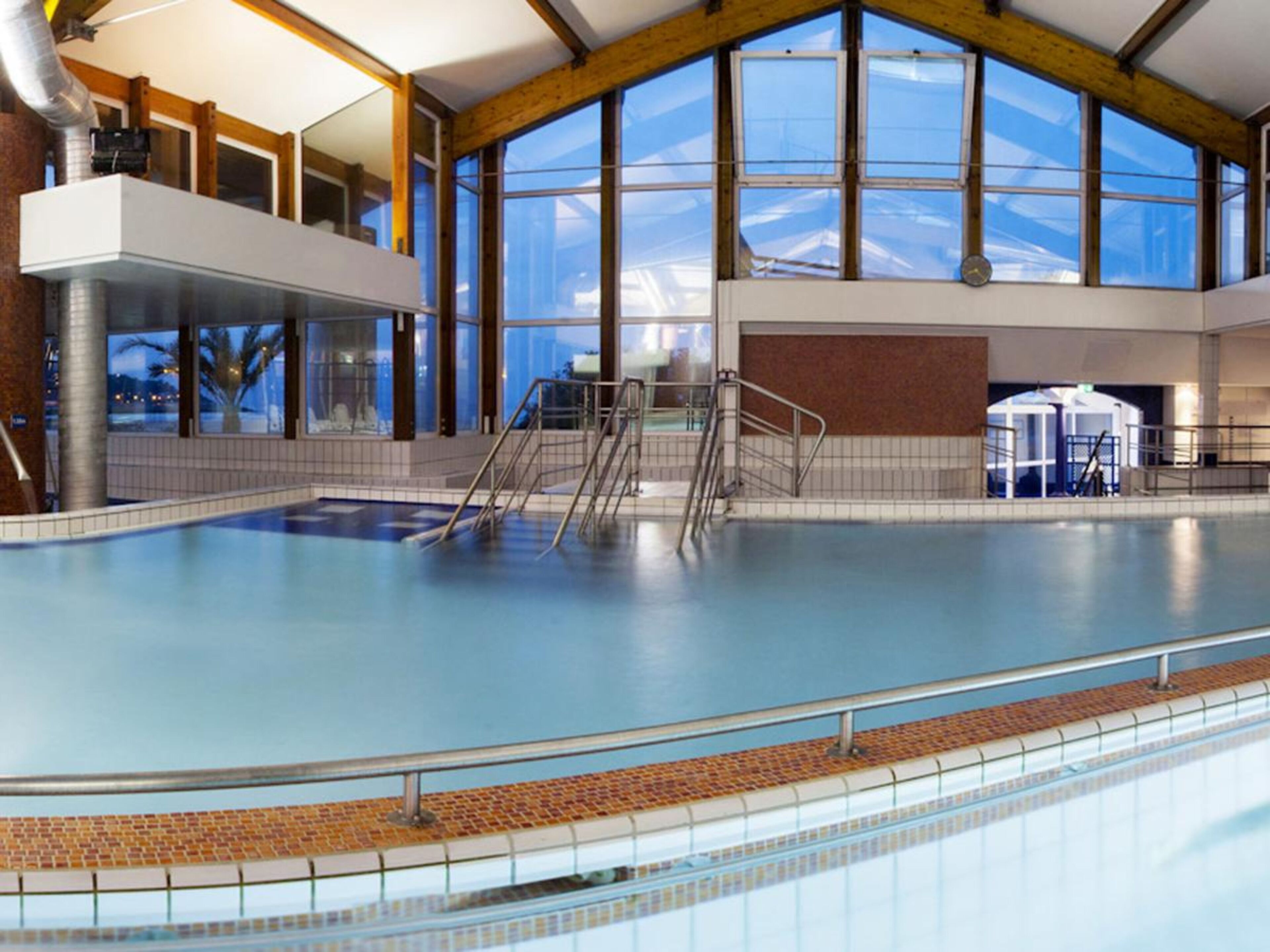 Une piscine intérieure dotée d’un haut plafond, de grandes fenêtres et d’un sol carrelé.