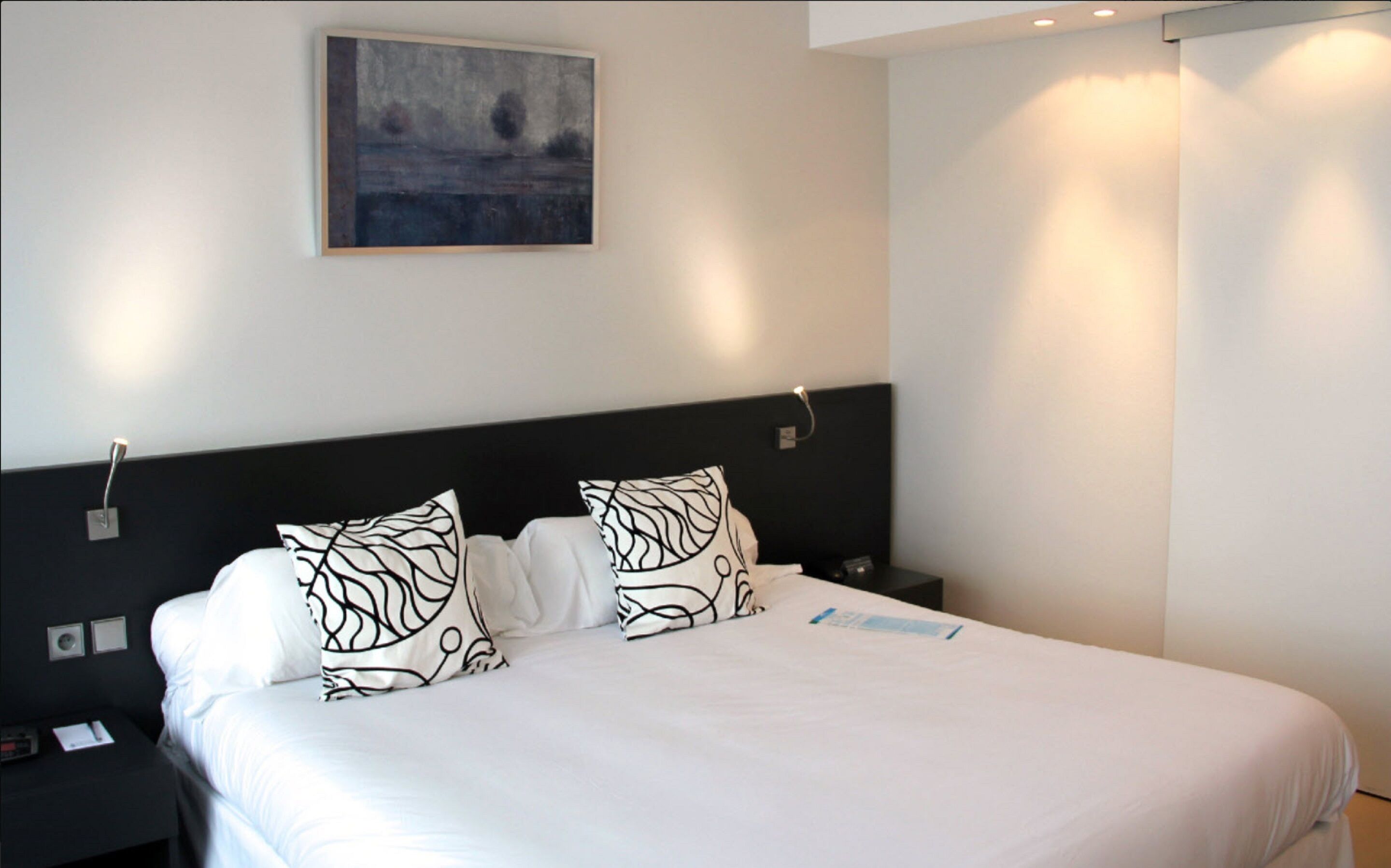 Una cama bien tendida con ropa blanca y almohadas estampadas en negro, un cabecero negro y un cuadro en la pared.