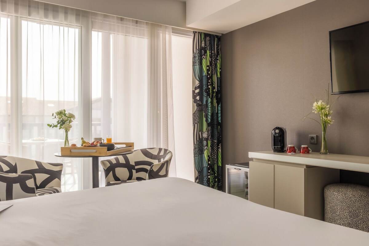Une chambre d’hôtel avec un lit, un bureau sur lequel se trouve un plateau de petit-déjeuner, une chaise, un téléviseur fixé au mur et une fenêtre avec des rideaux légers.
