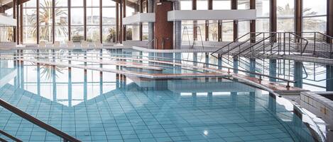 En indendørs swimmingpool med krystalklart blåt vand, omgivet af store vinduer med udsigt til palmer og et bakket landskab.