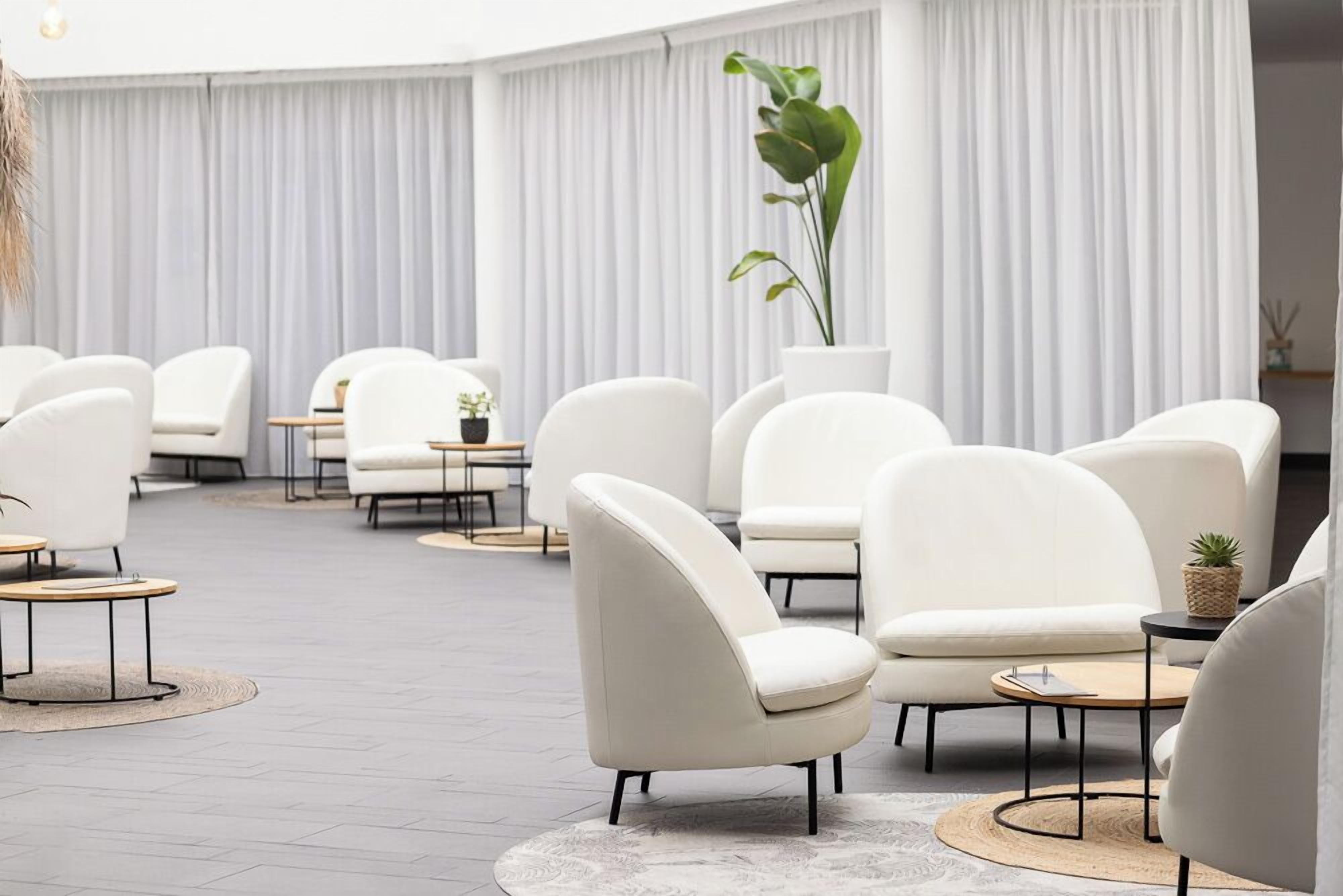 A modern lounge area with white leather chairs and round tables, featuring a large potted plant and a minimalist design.