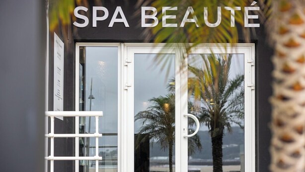 Eingangsbereich eines Spas mit Glastür, Palmen und Blick auf den Strand.