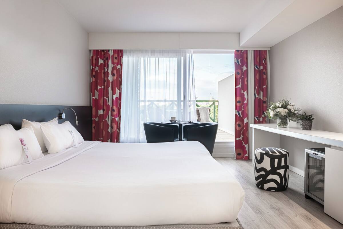 Une chambre d’hôtel comprenant un lit, un bureau, une chaise, une petite table et un balcon avec des rideaux.