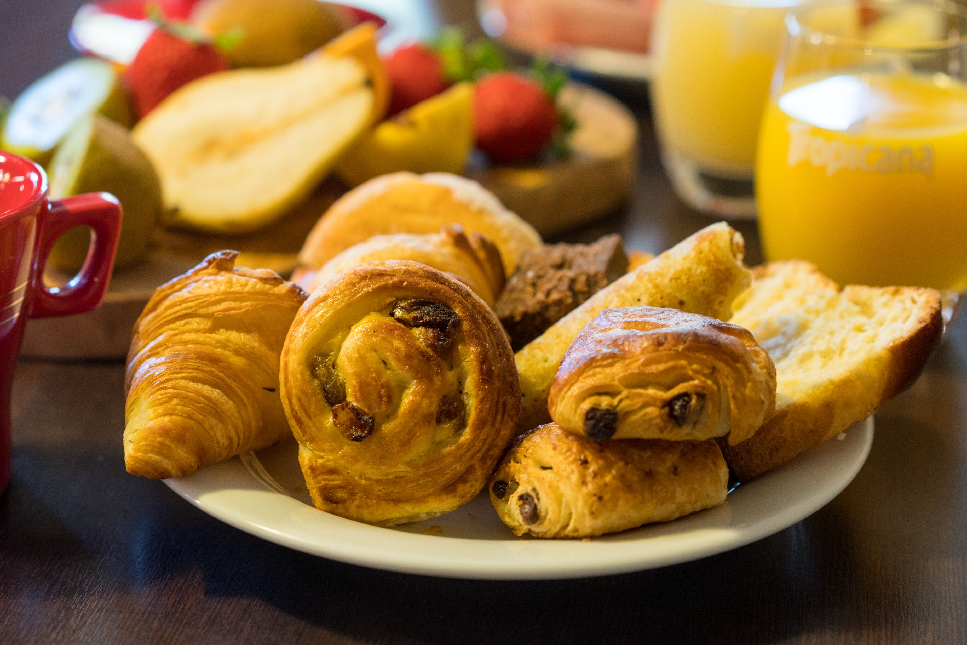 A plate of croissants and bread, a red mug, and a glass of orange juice on a table.