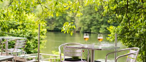 A table with two chairs and two glasses of wine, set against a backdrop of a lake and greenery.