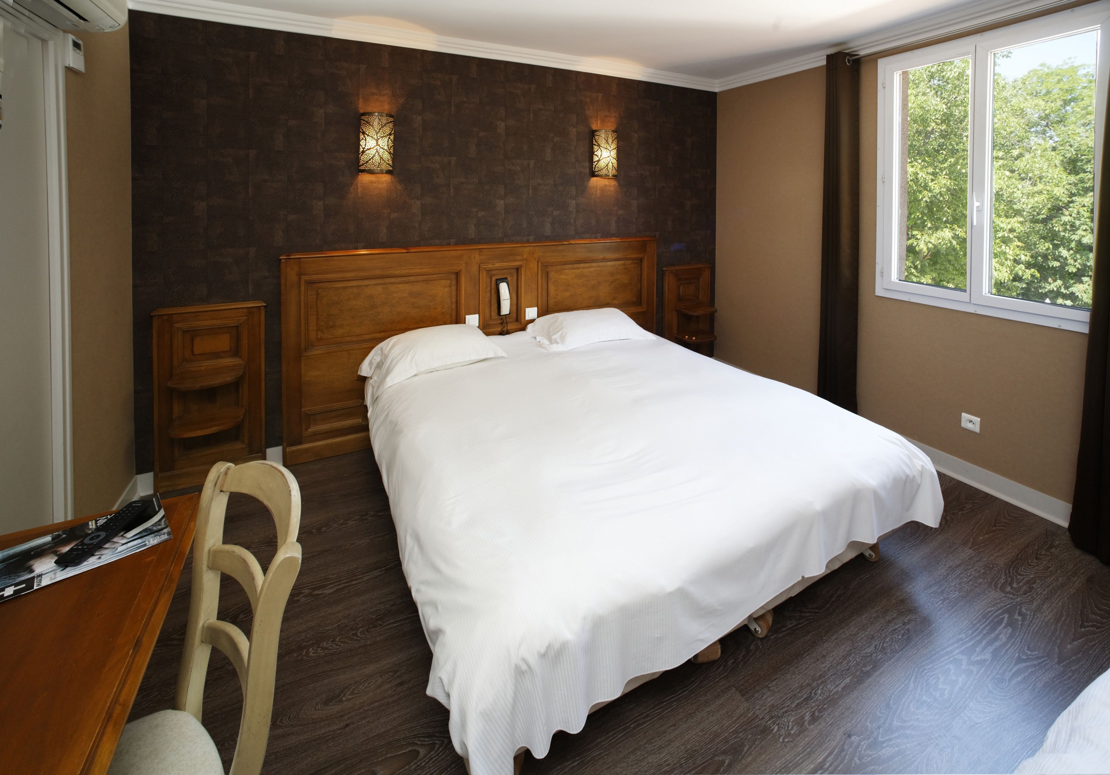 Una cama bien tendida en una habitación con piso de madera oscura, dos lámparas fijadas a la pared y un ventanal con cortinas.