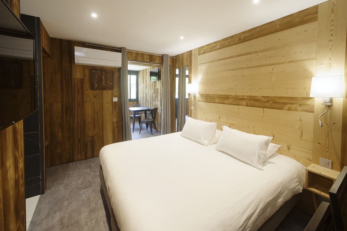 A bedroom with a wooden headboard, a bed with white linens, a bedside table with a lamp, and a view of a dining area through an open door.