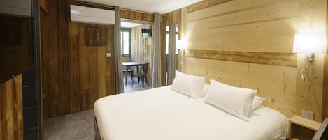 A bedroom with a wooden headboard, a bed with white linens, a bedside table with a lamp, and a view of a dining area through an open door.