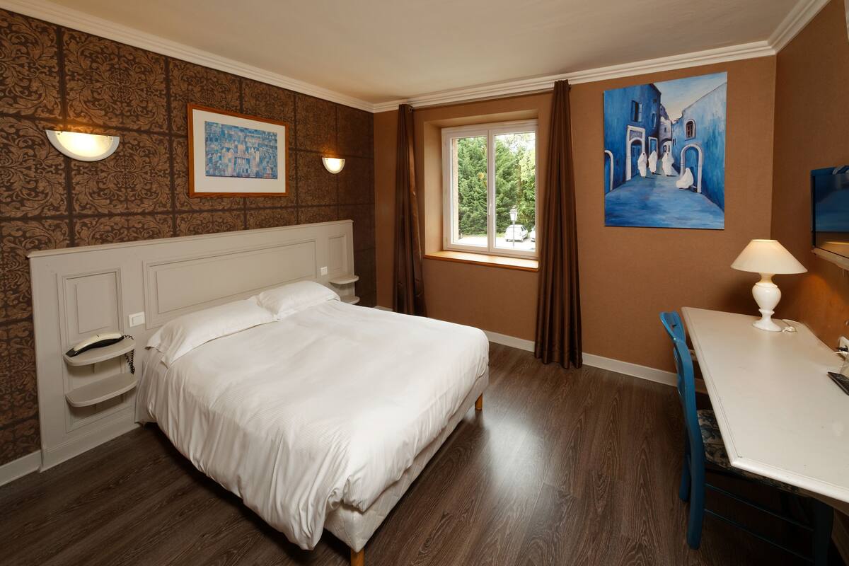 A bedroom with a bed, a desk, a chair, a window, and two paintings on the wall.