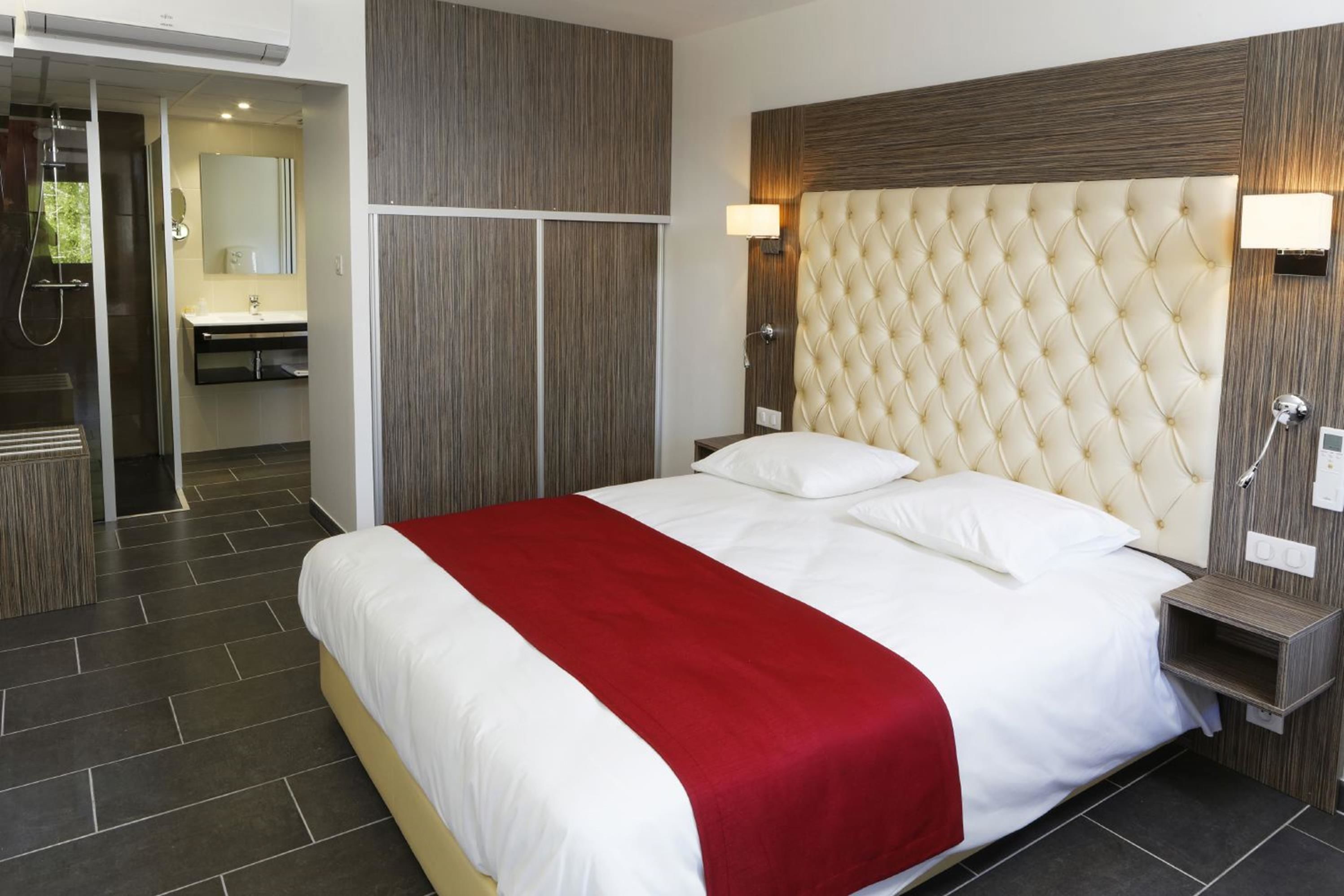 A hotel room with a large bed, a red throw, a wooden headboard, a nightstand, and a bathroom with a mirror and sink.