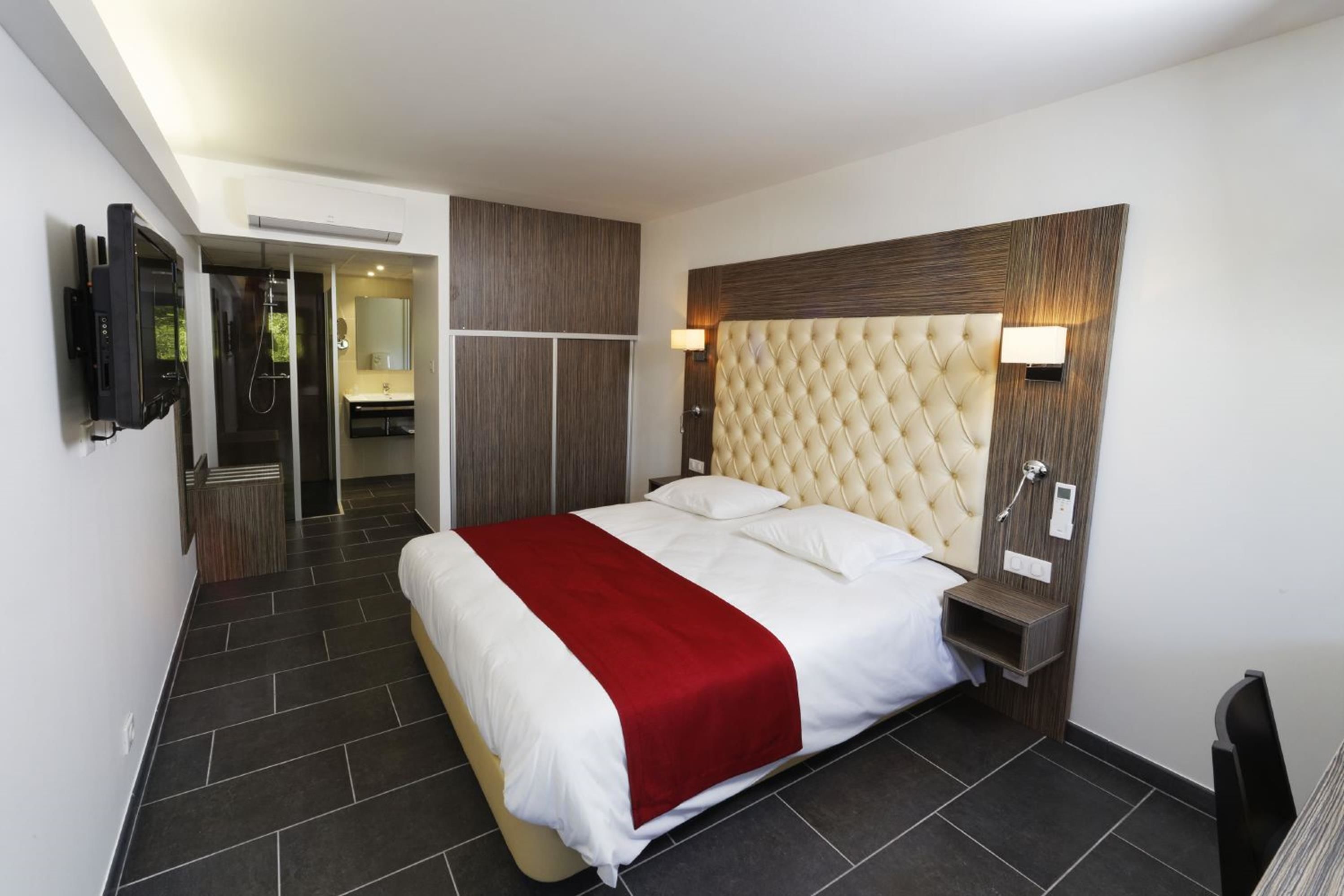 Quarto de hotel moderno com uma cama grande, televisão, banheiro e uma mesinha de cabeceira com abajur.