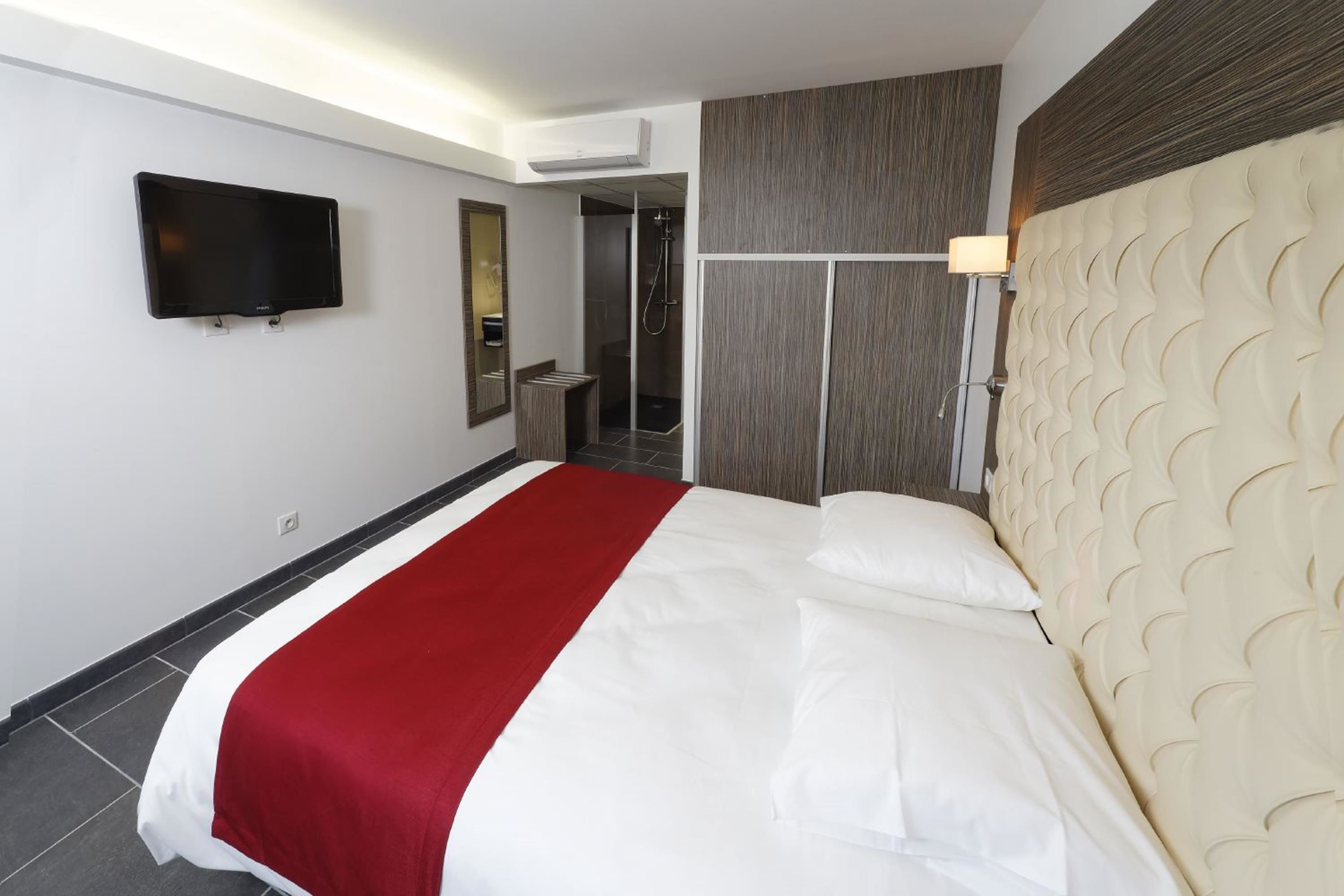 Camera d'albergo con un letto grande, una TV fissata al muro, cabina doccia e testiera imbottita con motivo decorativo.