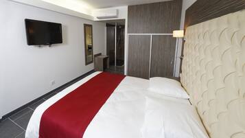 Camera d'albergo con un letto grande, una TV fissata al muro, cabina doccia e testiera imbottita con motivo decorativo.