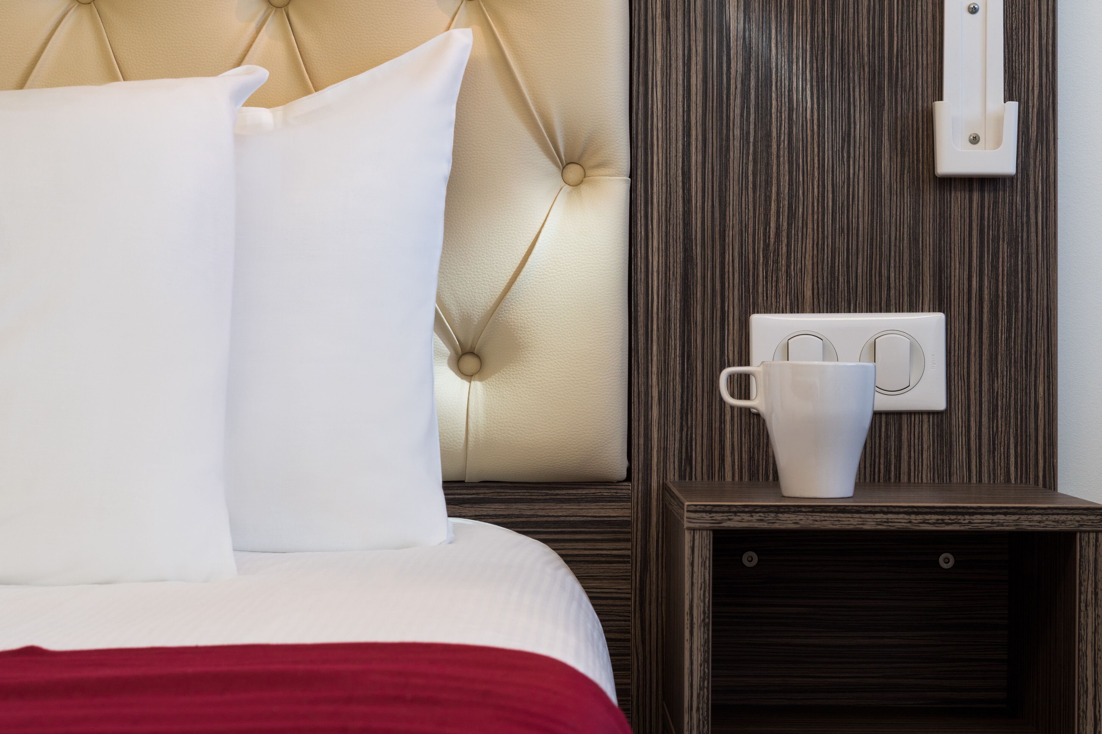 Cama bem arrumada com roupa de cama branca e vermelha, um travesseiro branco e uma mesa de cabeceira com uma caneca branca.