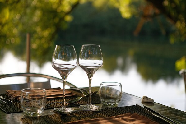 Una mesa con tres copas de vino y una taza pequeña, sobre un mantel tejido, con vista a un cuerpo de agua al fondo.