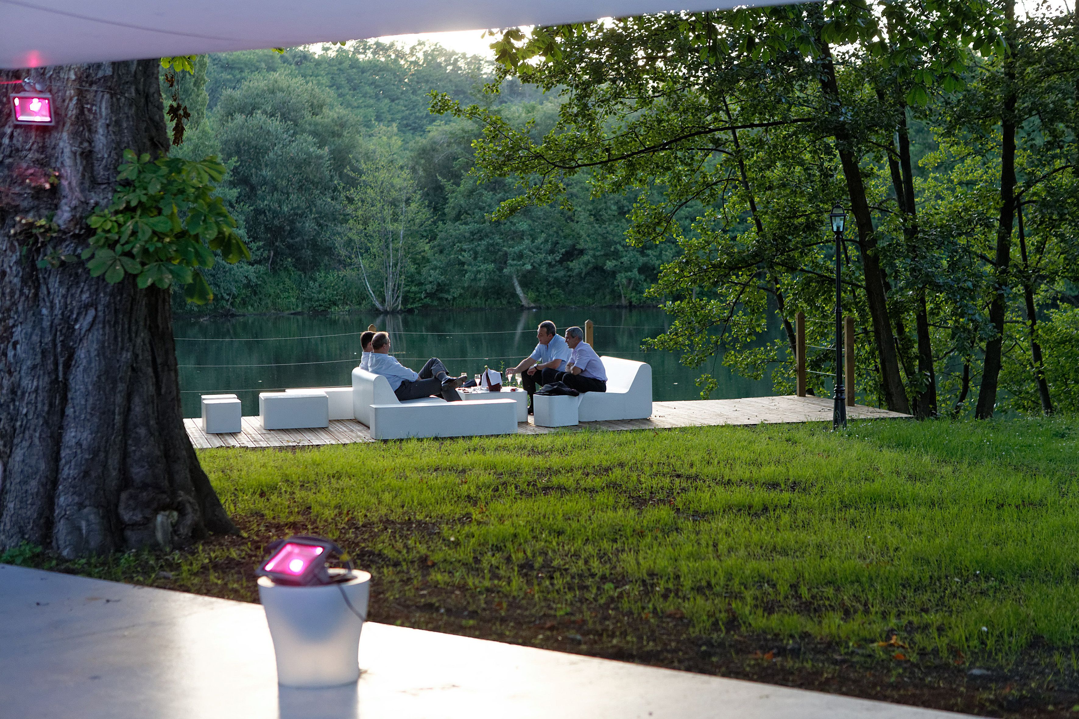 A group of people sitting on white outdoor sofas in a park-like setting with a tree and a fence in the background.