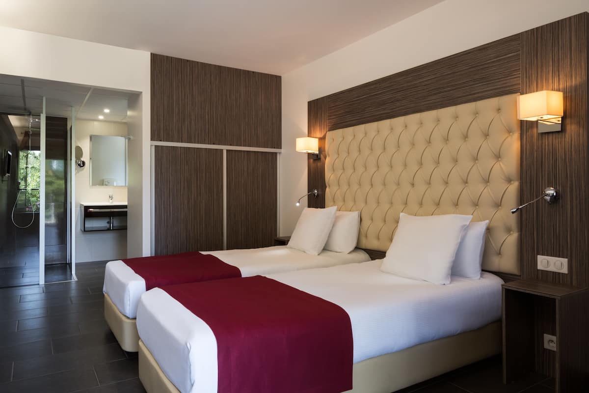A modern hotel room with two beds, a large tufted headboard, a glass shower area, and a wooden wall panel.