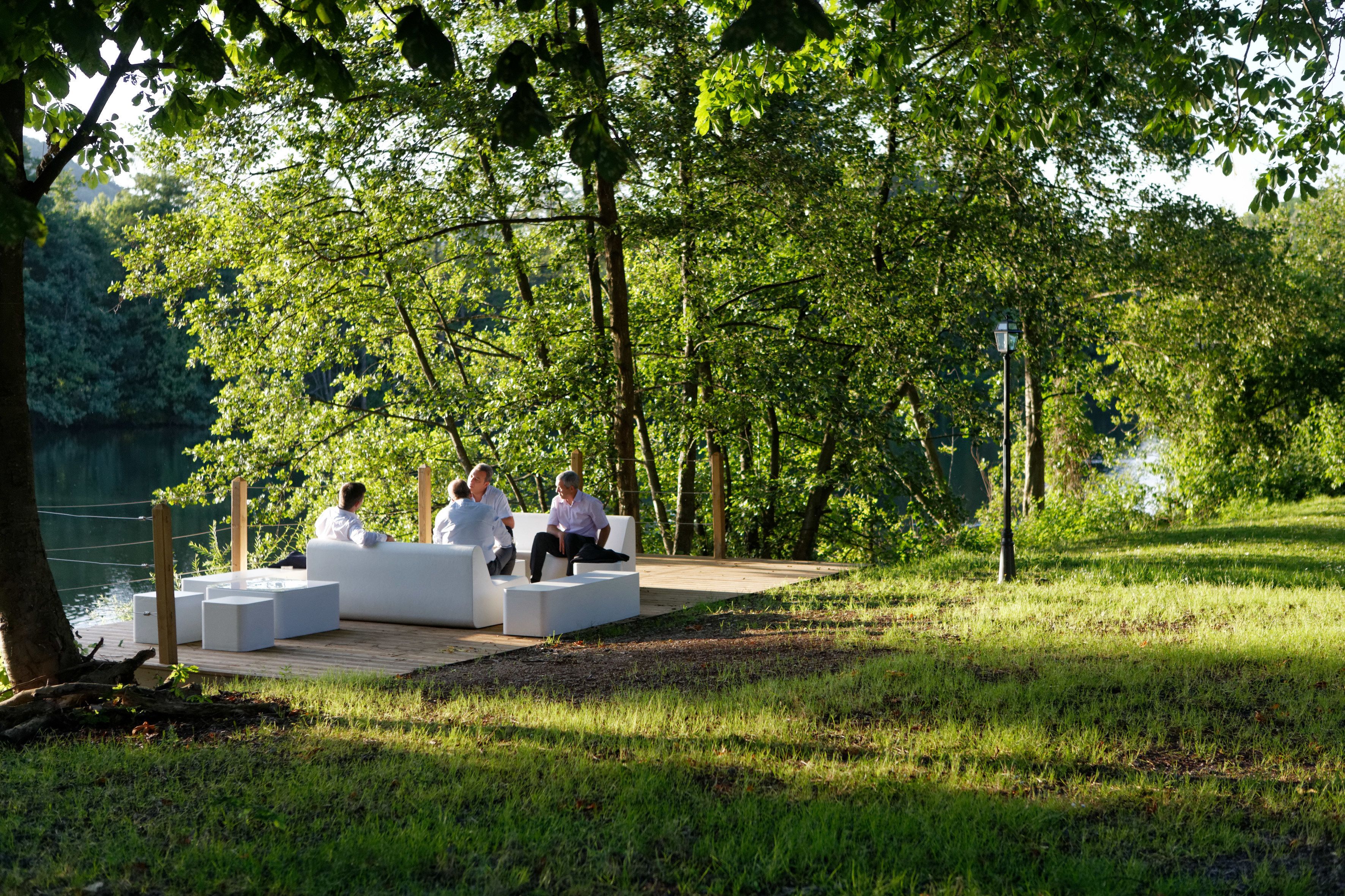 Un groupe de personnes est assis sur des meubles de jardin blancs dans un parc, avec un lac et des arbres en arrière-plan.