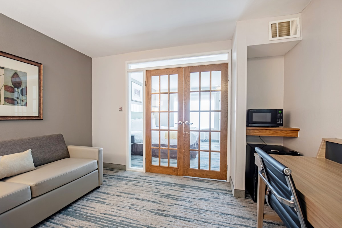 A modern hotel room with a sofa, a dining table, a microwave, and a large window with a view.