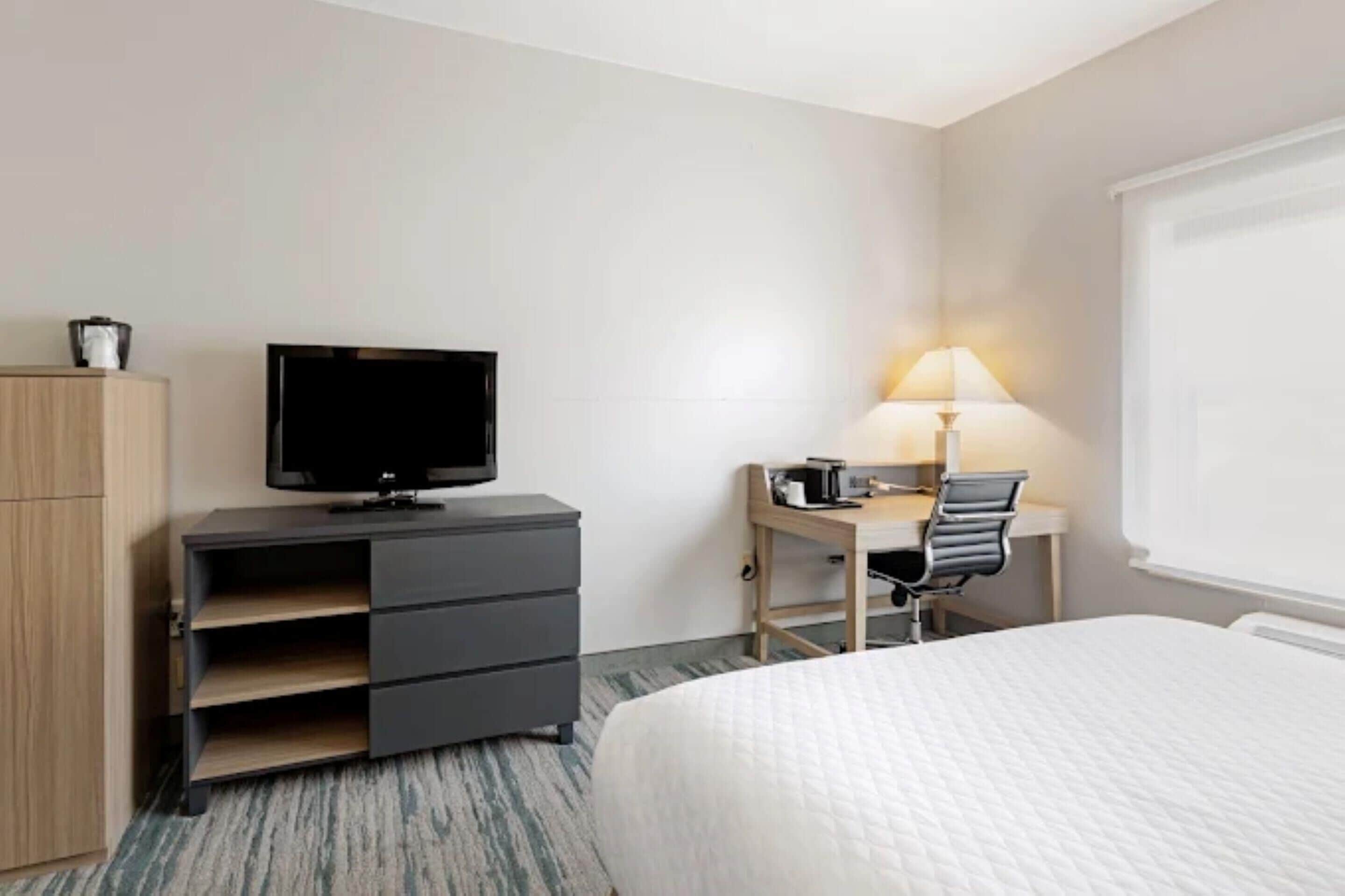 A hotel room with a bed, a desk with a chair, a television on a dresser, and a lamp.