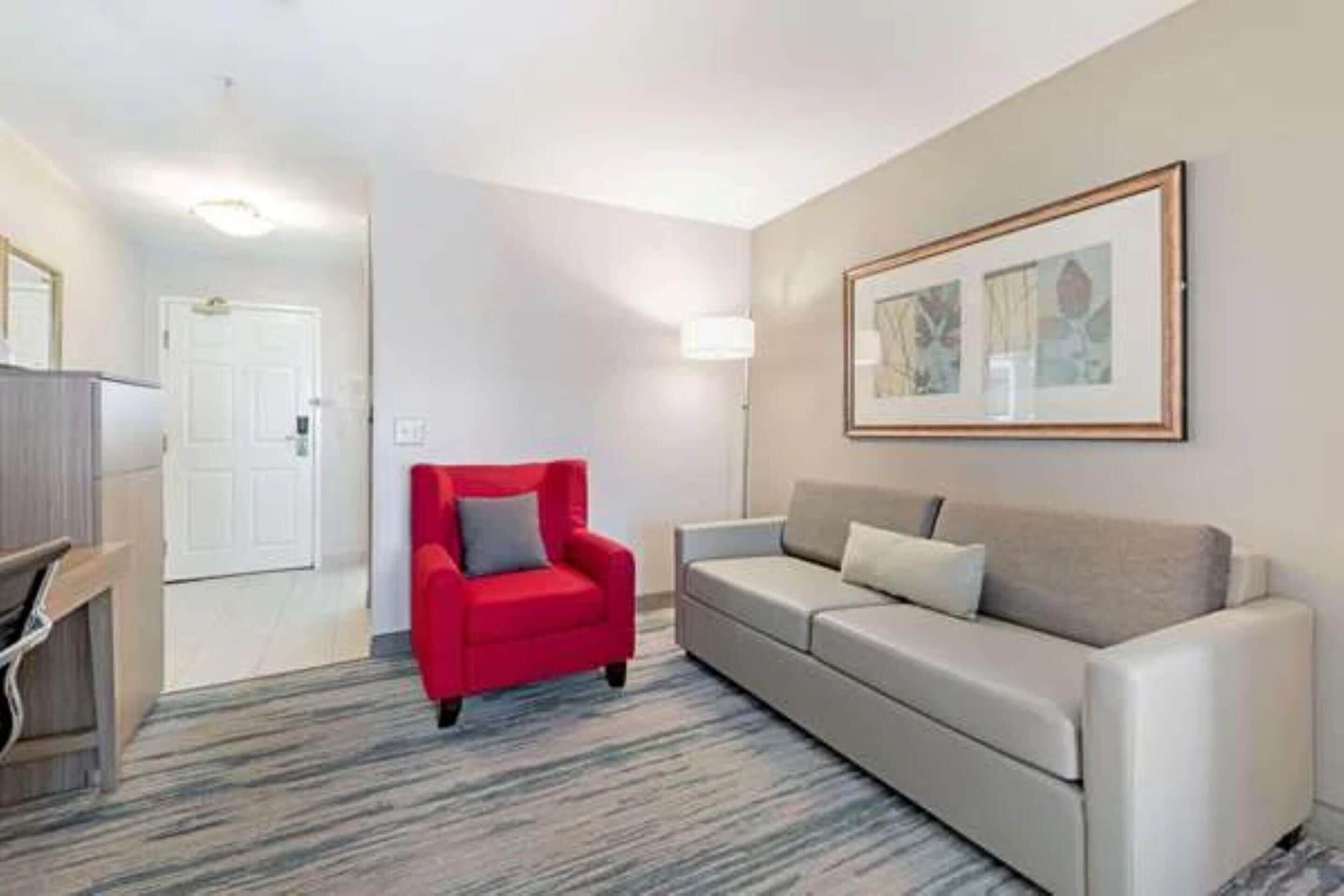 Une chambre d’hôtel avec un fauteuil rouge, un canapé beige, une œuvre d’art encadrée au mur et une porte donnant sur une autre pièce.