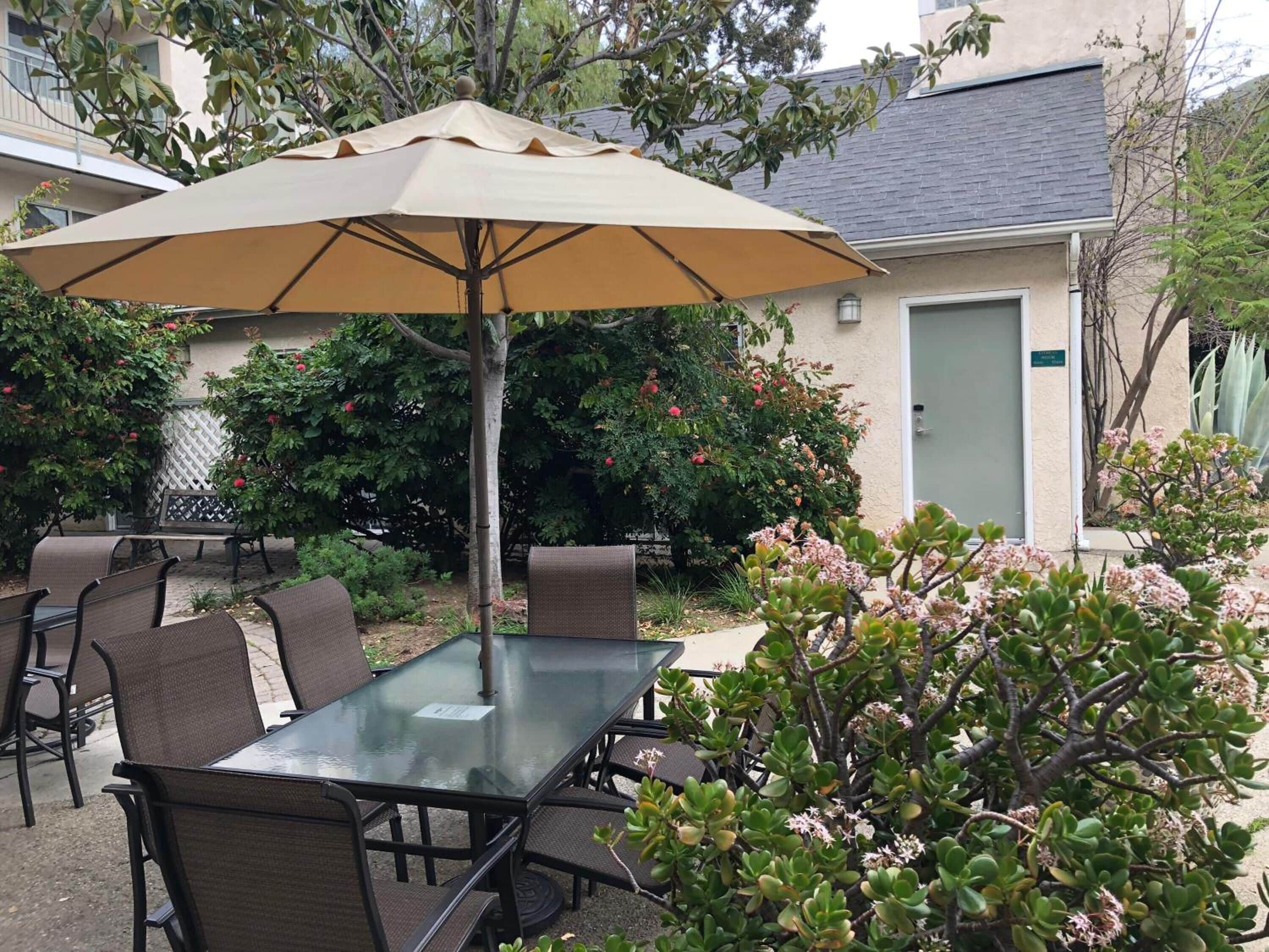 A glass-top table with chairs and a beige umbrella in a patio area with plants and a building in the background.