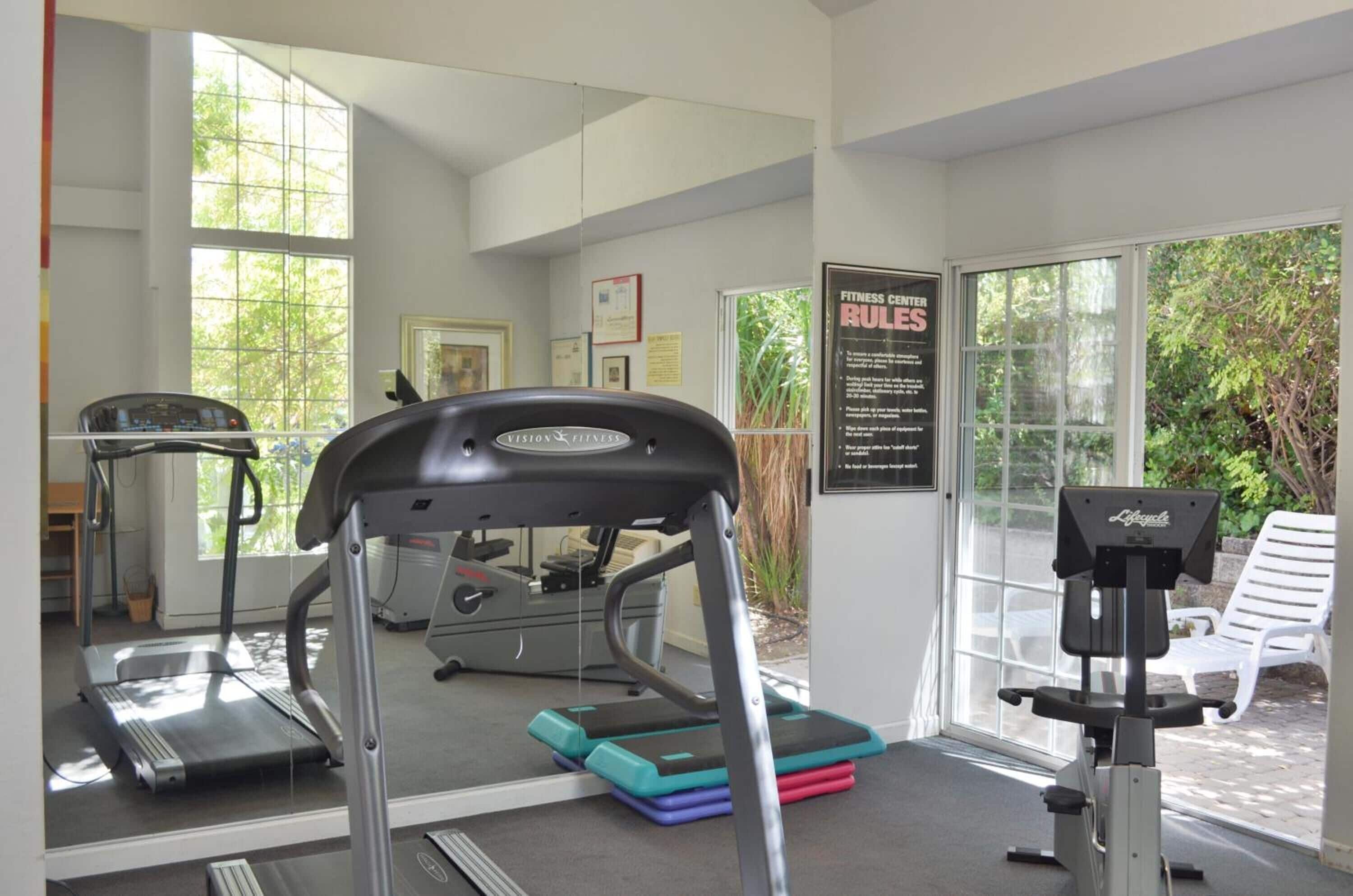 A gym with treadmills, a stationary bike, and a rowing machine. There are large windows and a door leading outside.