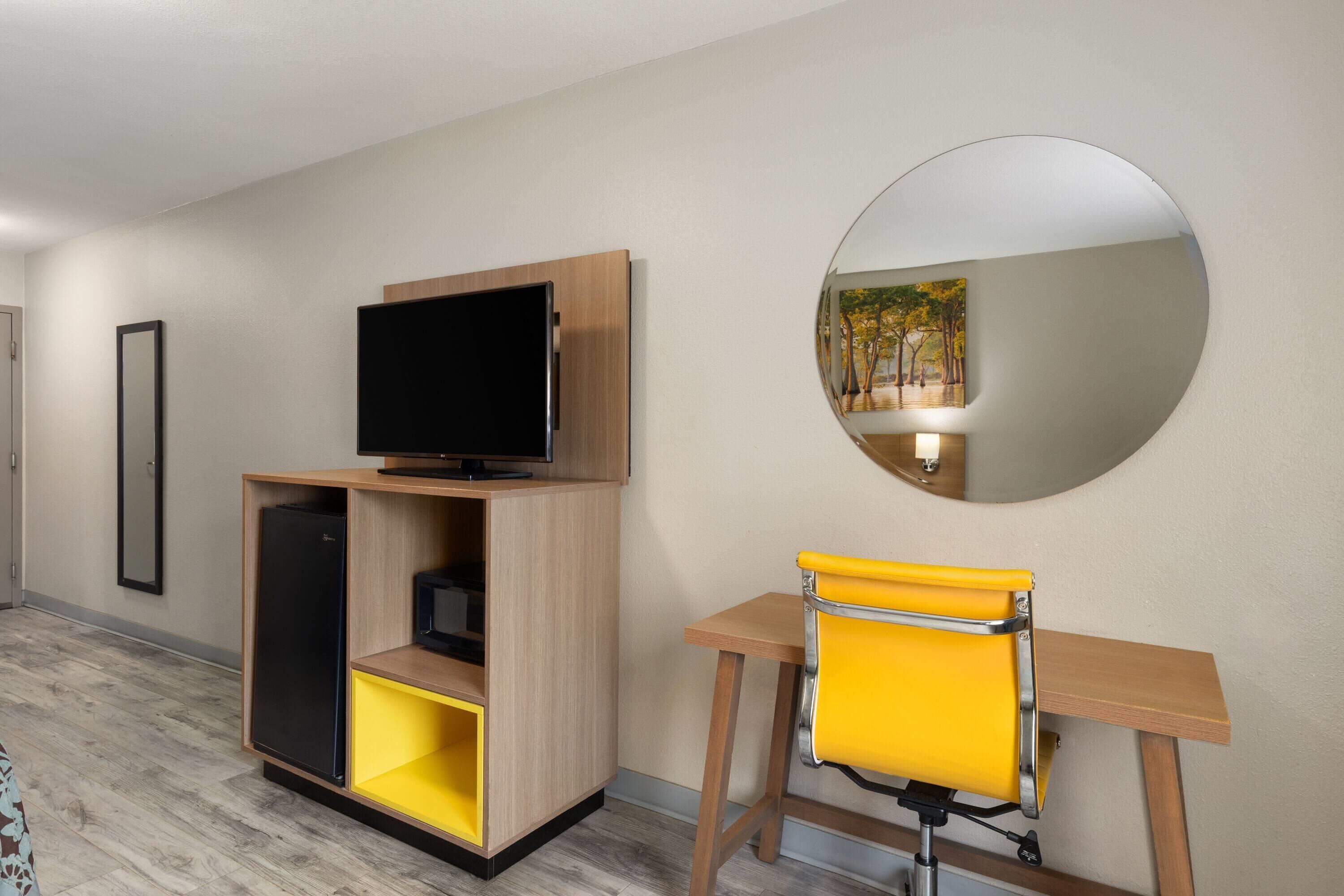 Une chambre d’hôtel avec un bureau en bois, une chaise jaune, un téléviseur à écran plat posé sur une commode en bois et un miroir rond accroché au mur.