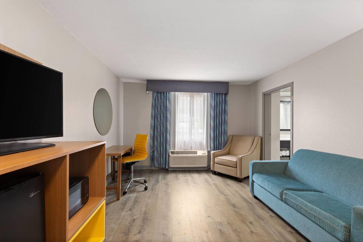 A hotel room with a flat-screen TV, a wooden entertainment unit, a yellow office chair, a teal sofa, and a large window with curtains.