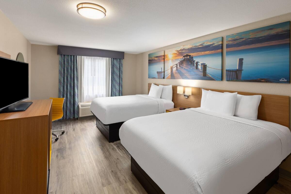 A hotel room with two beds, a desk, a TV, and a painting of a sunset over a pier.