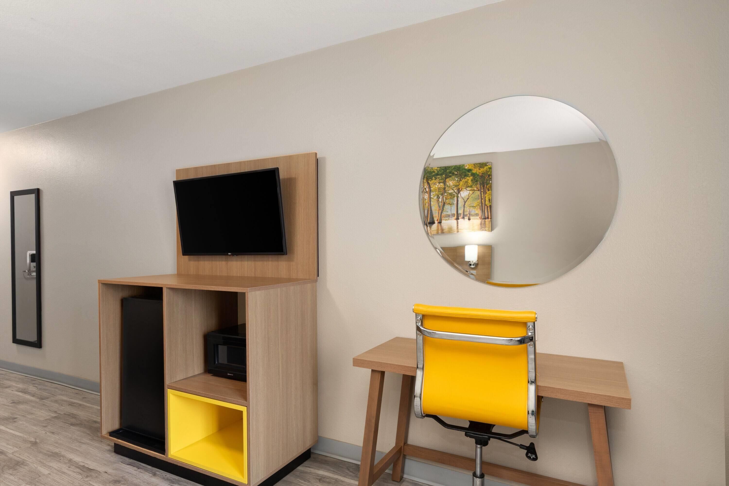 A hotel room with a TV mounted on a wooden shelf, a yellow chair, and a round mirror on the wall.