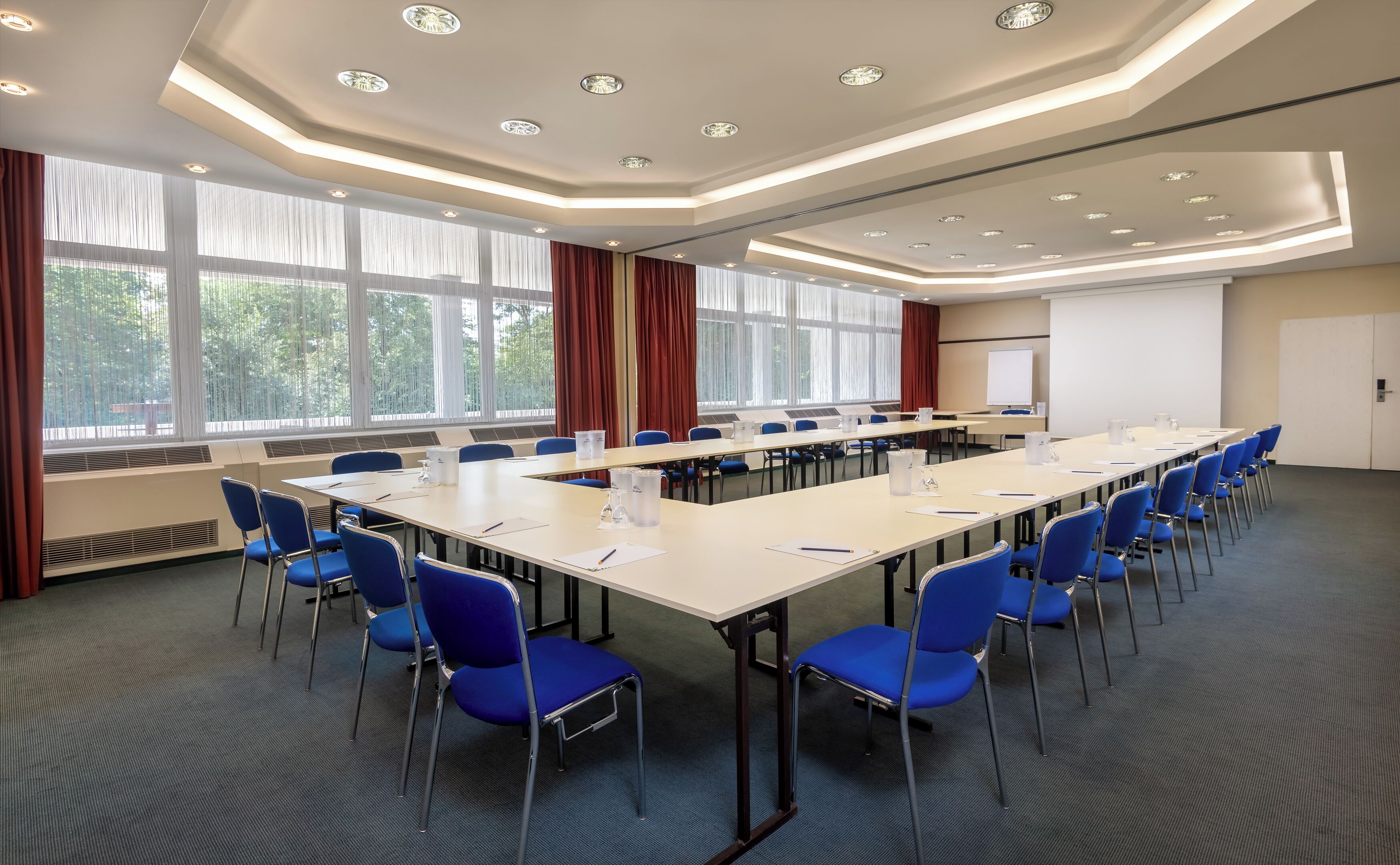 A conference room with long tables, blue chairs, and large windows with curtains.