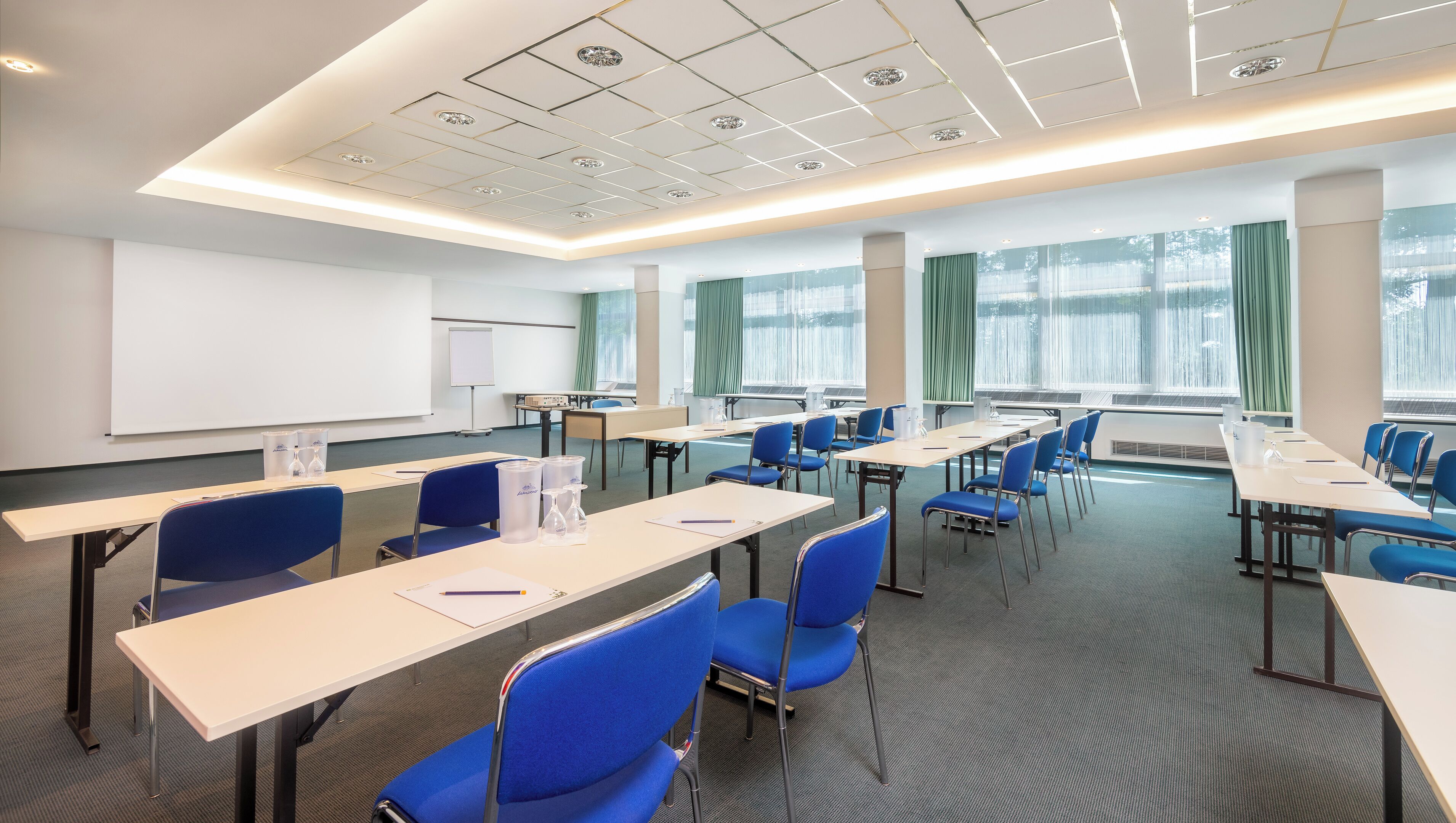 Un salón de conferencias con largas mesas, sillas azules, una pizarra y grandes ventanales con cortinas.