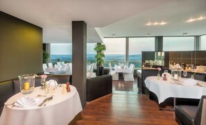 A restaurant with round tables, white tablecloths, and a view of the mountains. - Wyndham Garden Lahnstein Koblenz (Lahnstein)