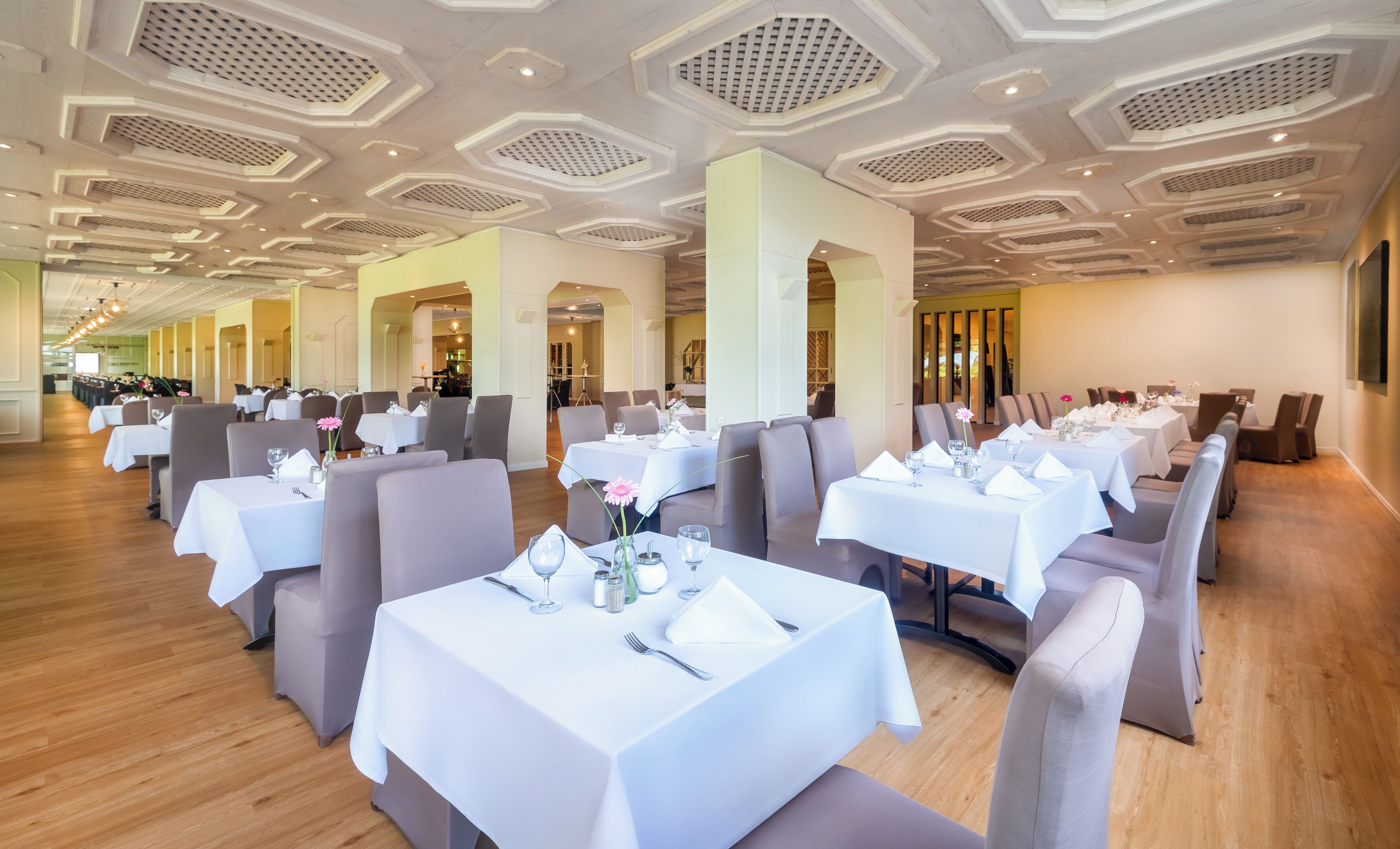 A restaurant with multiple tables set for dining, featuring a modern ceiling design and wooden flooring.