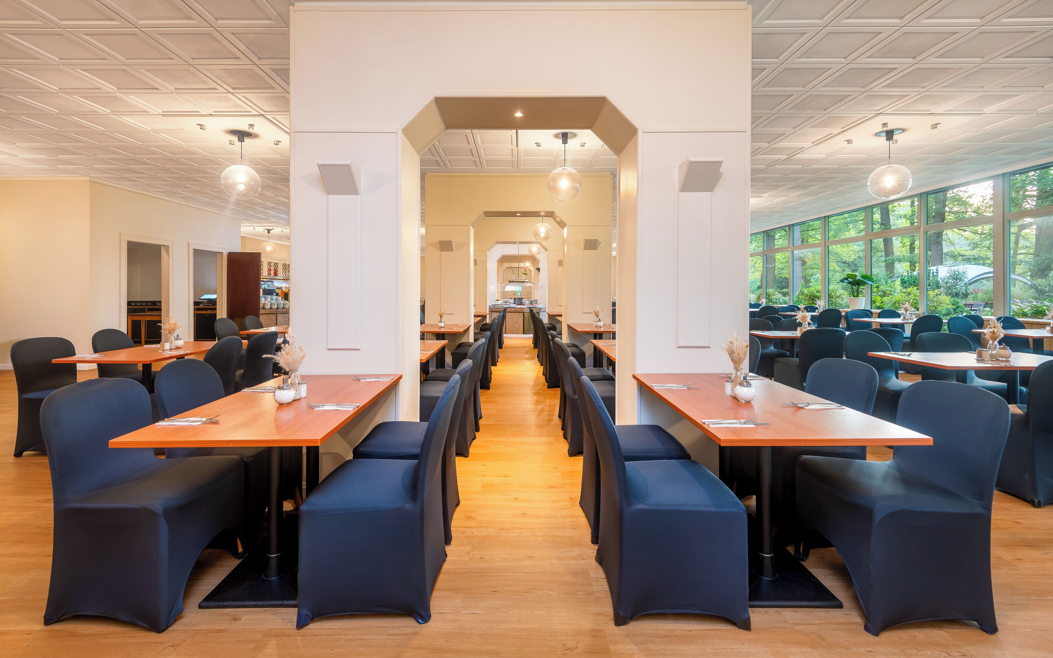 A spacious restaurant with long wooden tables and blue chairs, a central walkway, and large windows offering a view of greenery.